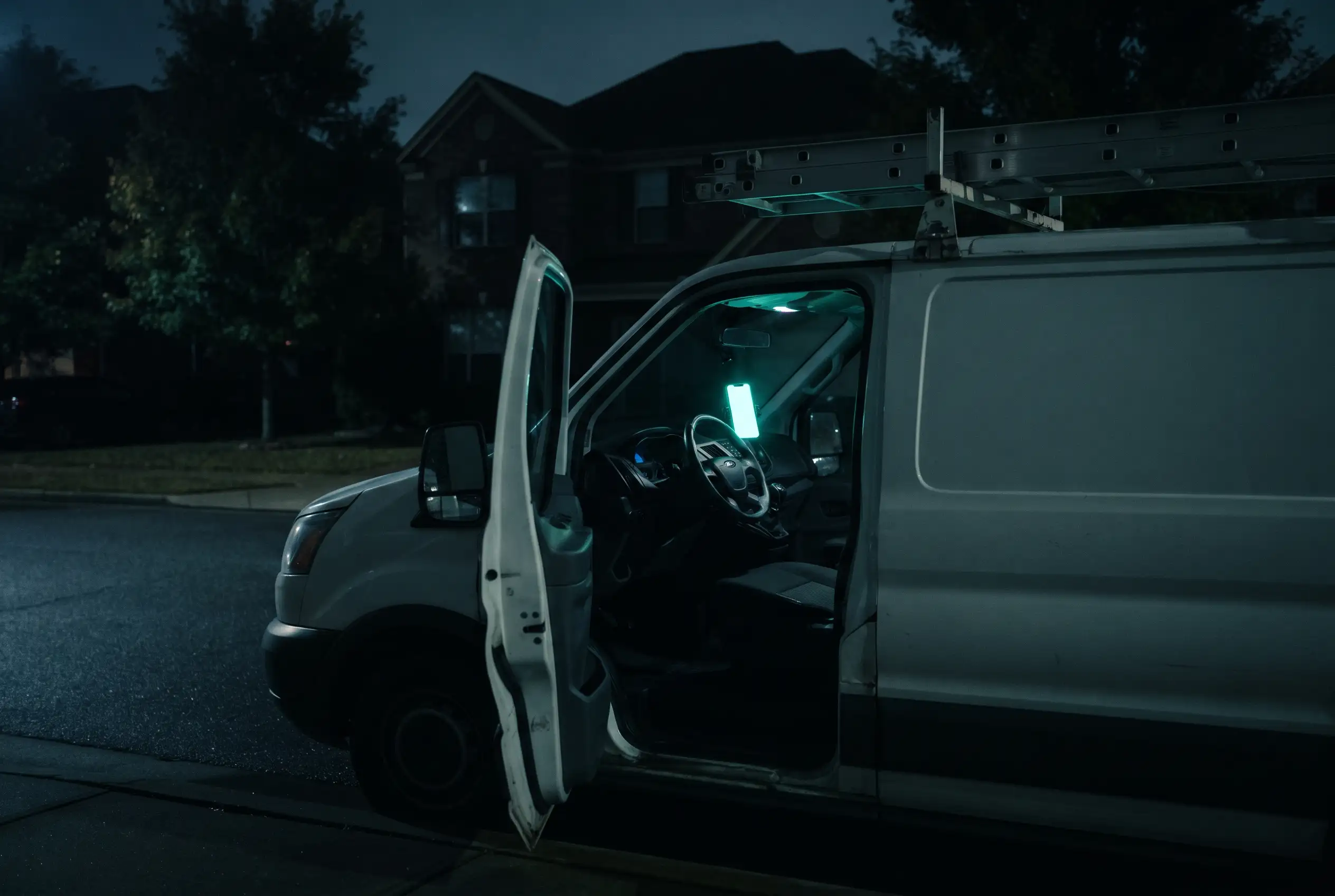 An HVAC contractor's van with a ladder rack outside a dark house at night, a phone glowing on the dashboard.
