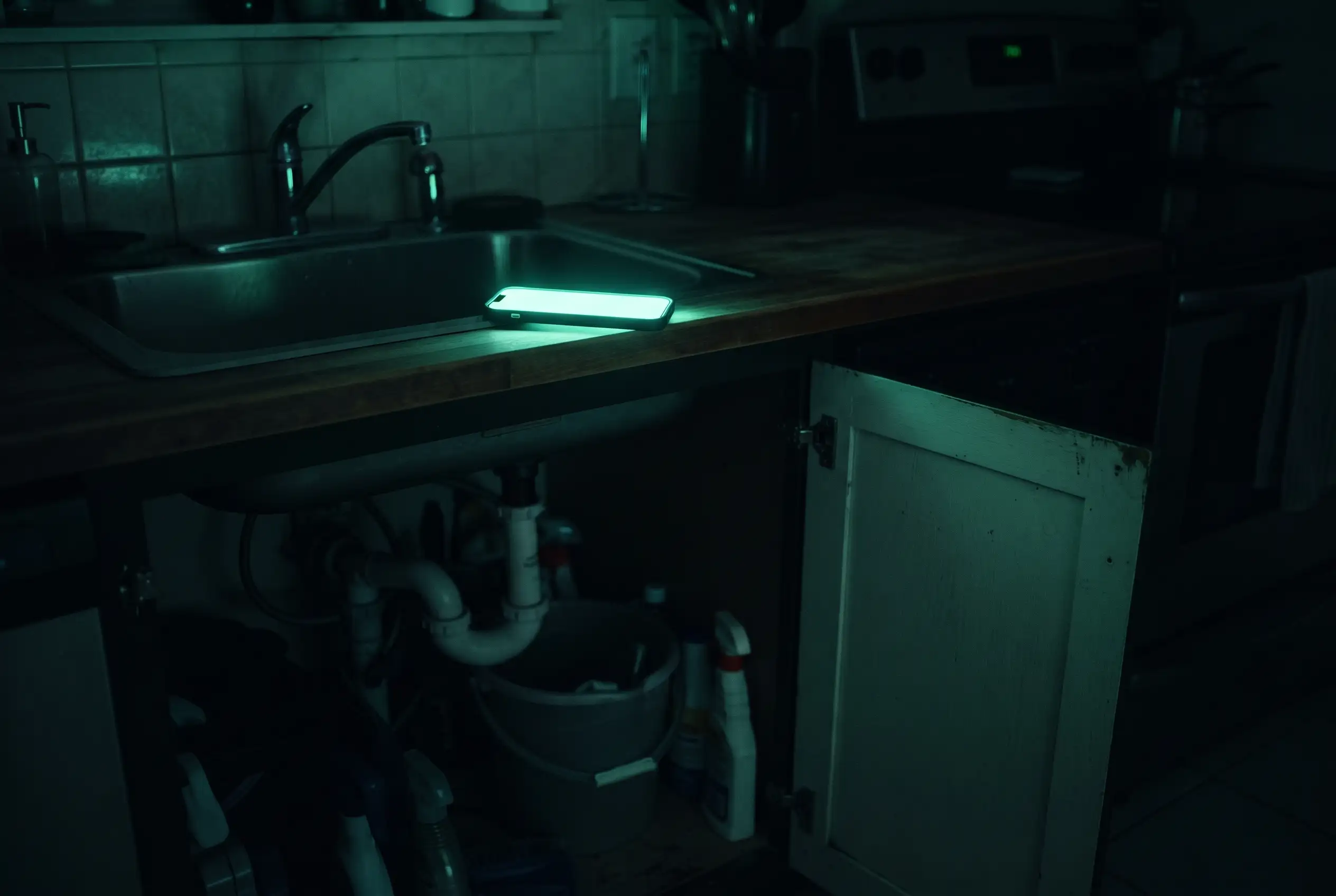 A dark kitchen at night, the cabinet under the sink open, a phone glowing on the counter above it.